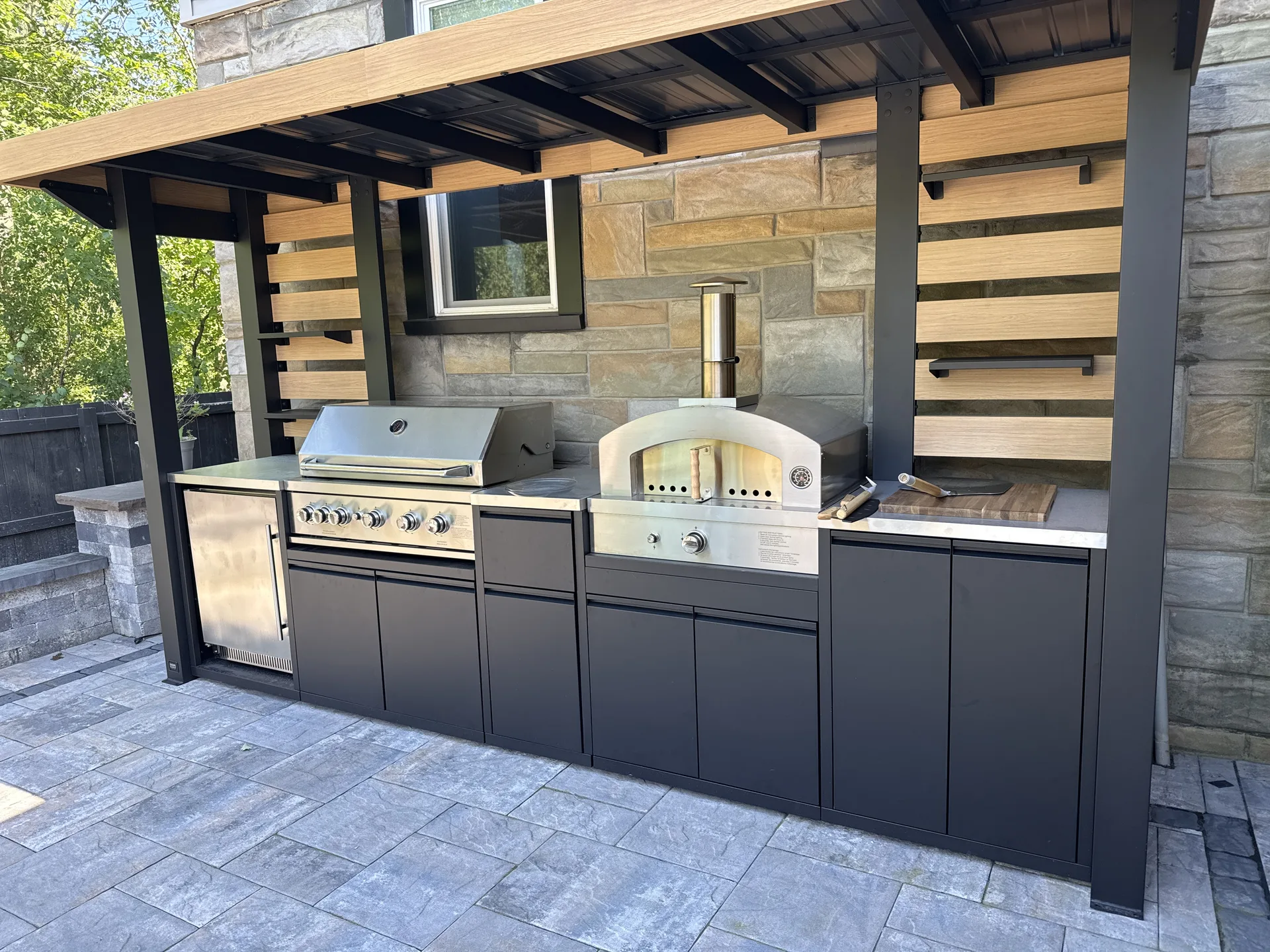 Outdoor grill and pizza oven beneath a timber roof with a slatted wood backdrop